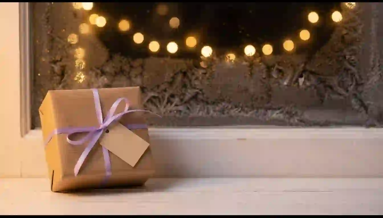 Diciembre es para calentar: el extra de Navidad hace que el cliente llegue a enero con ganas Caja de regalo envuelta en papel kraft con un lazo lavanda y una etiqueta en blanco, sobre el alféizar de una ventana con escarcha y luces de Navidad desenfocadas